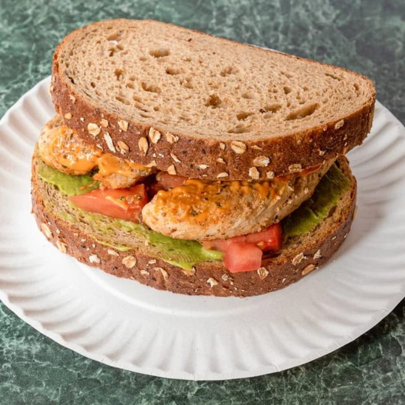 A chicken and avocado sandwich with tomatoes and sauce on whole grain bread, served on a paper plate.